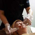 Operatrice di Manipura che applica un prodotto esfoliante sul viso di una cliente durante un trattamento professionale, indossando mascherina e guanti.