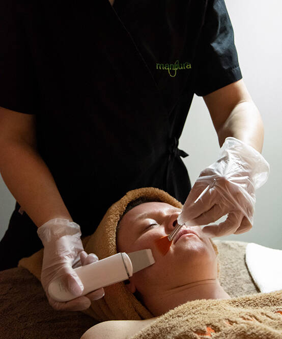 Operatrice di Manipura che applica un prodotto esfoliante sul viso di una cliente durante un trattamento professionale, indossando mascherina e guanti.