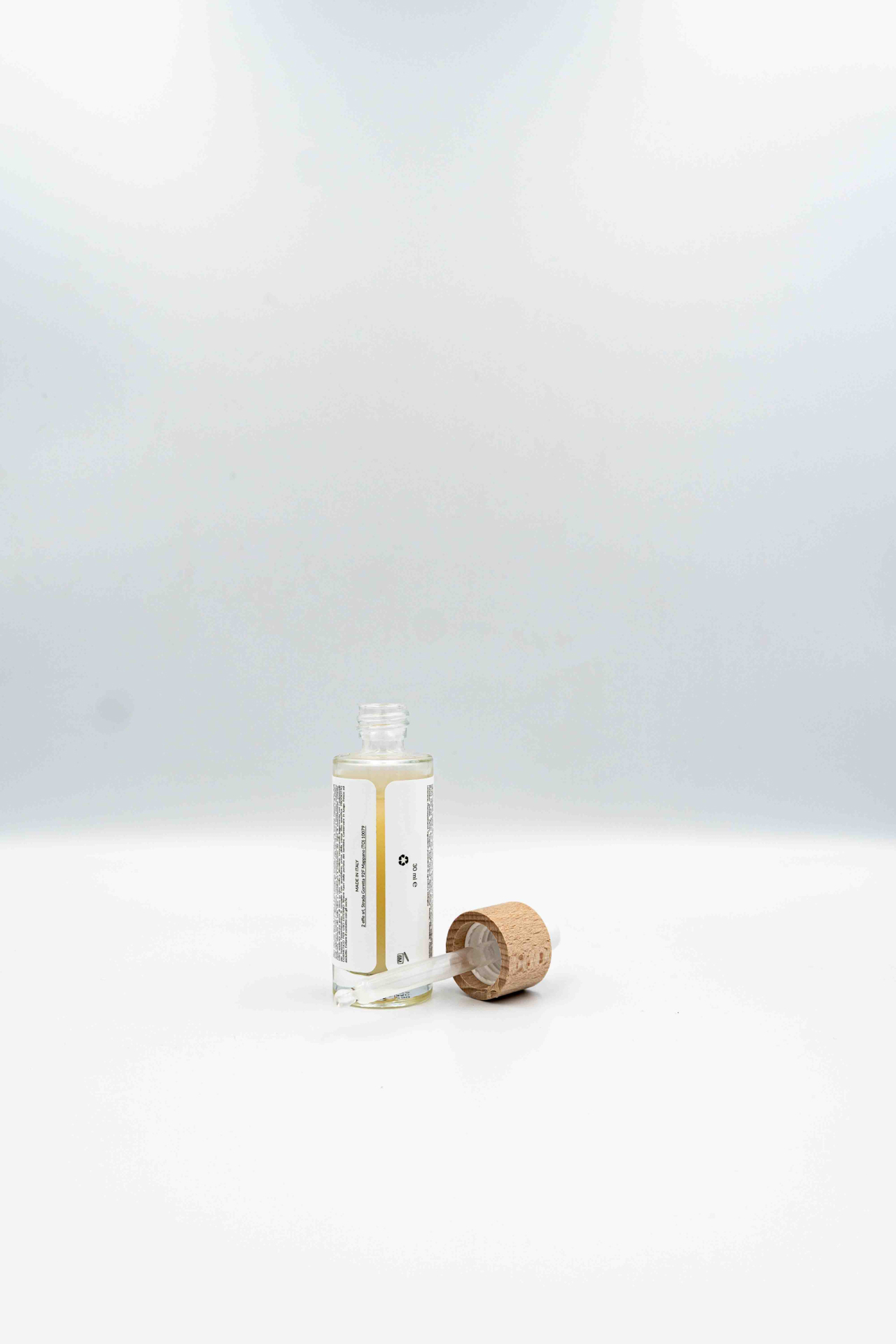 Flacone aperto di siero con etichetta retro visibile e contagocce appoggiato accanto al tappo in legno, su sfondo bianco.