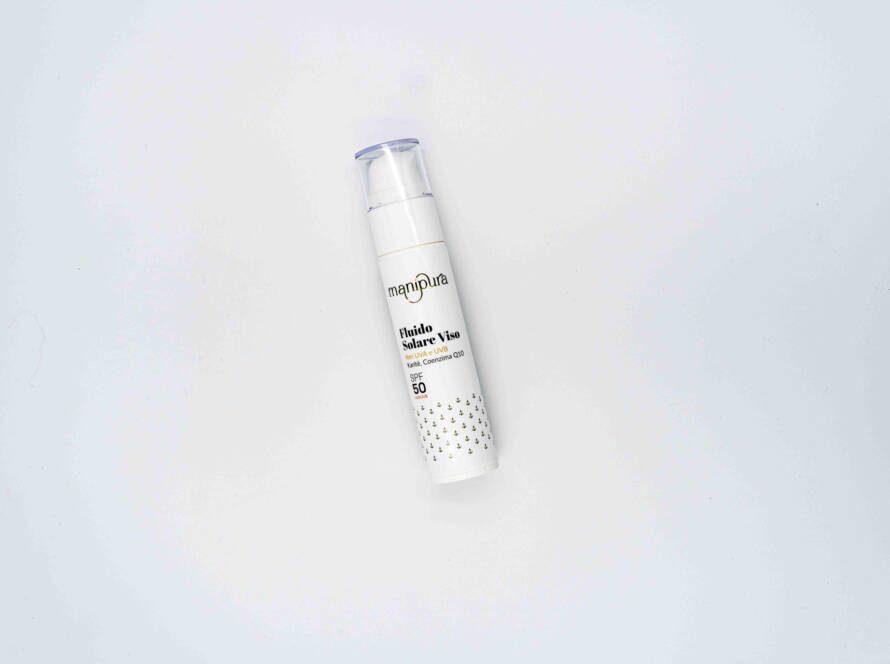 Flacone del Fluido Solare Viso SPF 50 di Manipura, fotografato dall’alto su sfondo bianco, con tappo trasparente e packaging bianco decorato.