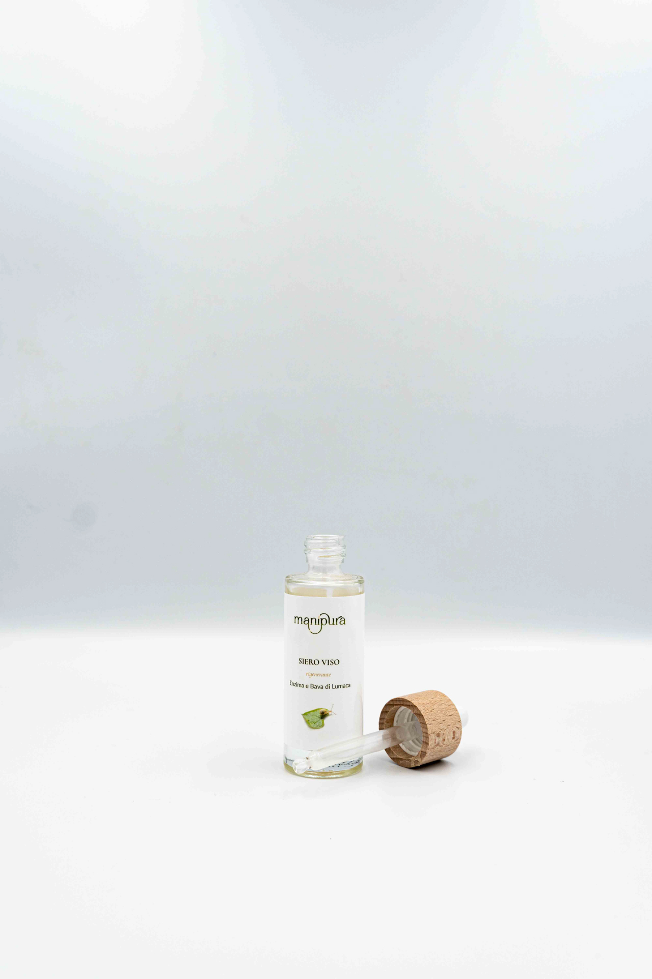 Flacone aperto di siero viso rigenerante Manipura alla bava di lumaca con contagocce e tappo in legno appoggiati su sfondo bianco.